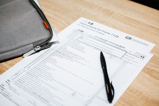 A tax form labeled Form 1120 sits on a wooden table. A black pen rests on the form, and a gray zippered pouch is partially visible beside it.