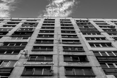 A tall, multi-story residential building is viewed from below, emphasizing its height and structure. The facade is worn, with visible satellite dishes attached to the walls. The windows are arranged in a repetitive pattern typical of high-rise apartments. The sky is partially visible, showing some scattered clouds.