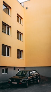 A black car is parked in front of an apricot-colored building with multiple windows. The building has a modern facade, with windows featuring dark trim. The lighting suggests either early morning or late afternoon, casting a soft glow on the scene. The street is relatively empty and the building appears to be residential.