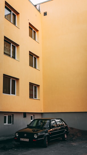 A black car is parked in front of an apricot-colored building with multiple windows. The building has a modern facade, with windows featuring dark trim. The lighting suggests either early morning or late afternoon, casting a soft glow on the scene. The street is relatively empty and the building appears to be residential.