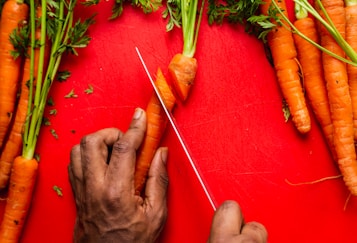 A hand expertly slices a fresh carrot on a vivid red cutting board. Several whole carrots with green tops are placed beside, creating a vibrant contrast. The angle and focus suggest culinary preparation.