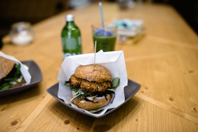 A close-up view of a gourmet burger served on a square plate with a wooden skewer holding it together. Accompanying the burger is another burger partially visible on the left side. A green bottled drink and a glass with a blue straw containing a green beverage are in the background, suggesting a casual dining setting on a wooden table.