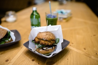 A close-up view of a gourmet burger served on a square plate with a wooden skewer holding it together. Accompanying the burger is another burger partially visible on the left side. A green bottled drink and a glass with a blue straw containing a green beverage are in the background, suggesting a casual dining setting on a wooden table.