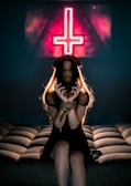 A woman with long hair sits on a cushioned surface with her hands bound by rope. She is wearing a dark dress and a headpiece resembling spikes. In the background, a neon sign with an inverted cross is prominently displayed, casting a reddish glow.