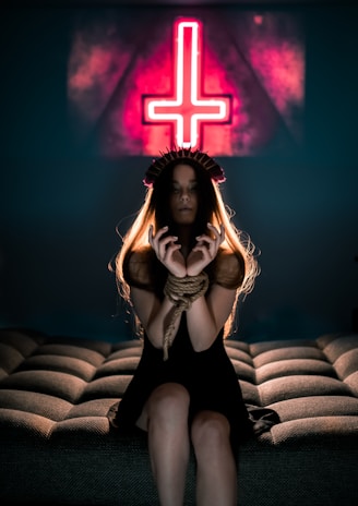 A woman with long hair sits on a cushioned surface with her hands bound by rope. She is wearing a dark dress and a headpiece resembling spikes. In the background, a neon sign with an inverted cross is prominently displayed, casting a reddish glow.