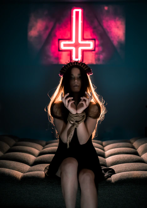 A woman with long hair sits on a cushioned surface with her hands bound by rope. She is wearing a dark dress and a headpiece resembling spikes. In the background, a neon sign with an inverted cross is prominently displayed, casting a reddish glow.