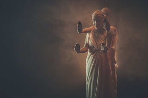 A woman in a white robe is centered, with four arms extending outward, each raised in various mudras or gestures. Another person partially obscured stands closely behind her, creating a layered visual effect in a smoky, dimly lit environment.