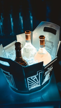 A well-lit bucket with intricate geometric designs holds three bottles, partially submerged in ice. The bottles have metallic caps and labels, with one being darker than the others. The setting appears to be stylish and modern, with a dimly lit background emphasizing the bucket as the focal point.