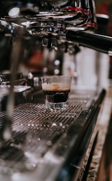 A freshly brewed espresso in a glass sits on the metallic tray of a coffee machine. The machine is prominently featured with its various shiny components reflecting light. The setting suggests a café environment, with the focus on the rich crema topping the dark coffee.