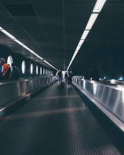 An elongated moving walkway inside a tunnel-like structure with round windows providing soft natural light. Several people are seen in the distance, walking along the travelator, with their backs turned, heading towards the brighter end of the passage. The ceiling has a grid pattern with fluorescent lighting fixtures.