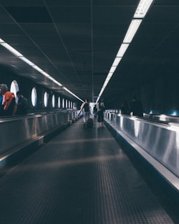 An elongated moving walkway inside a tunnel-like structure with round windows providing soft natural light. Several people are seen in the distance, walking along the travelator, with their backs turned, heading towards the brighter end of the passage. The ceiling has a grid pattern with fluorescent lighting fixtures.