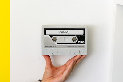 A hand is holding a grey rectangular object resembling a cassette tape against a white background. The cassette has markings including the word 'MARK' at the top, 'C90' on the right, and 'SUPER QUALITY LOW NOISE' at the bottom. A portion of the left edge shows a bright yellow color.