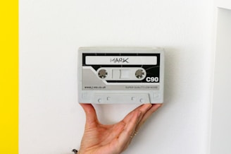A hand is holding a grey rectangular object resembling a cassette tape against a white background. The cassette has markings including the word 'MARK' at the top, 'C90' on the right, and 'SUPER QUALITY LOW NOISE' at the bottom. A portion of the left edge shows a bright yellow color.