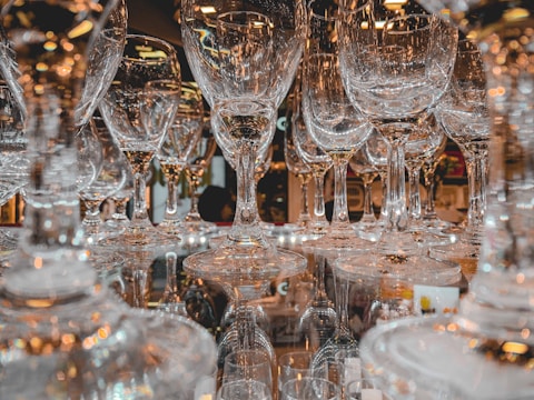 A collection of clear wine glasses is clustered together, creating an intricate pattern and reflecting light in various directions. The glasses are arranged on a reflective surface, enhancing the visual complexity.