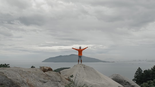 man in red jacket standing on gray rock formation under white clouds during daytime in Hải Vân Pass Vietnam