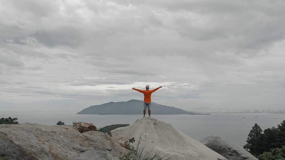 man in red jacket standing on gray rock formation under white clouds during daytime