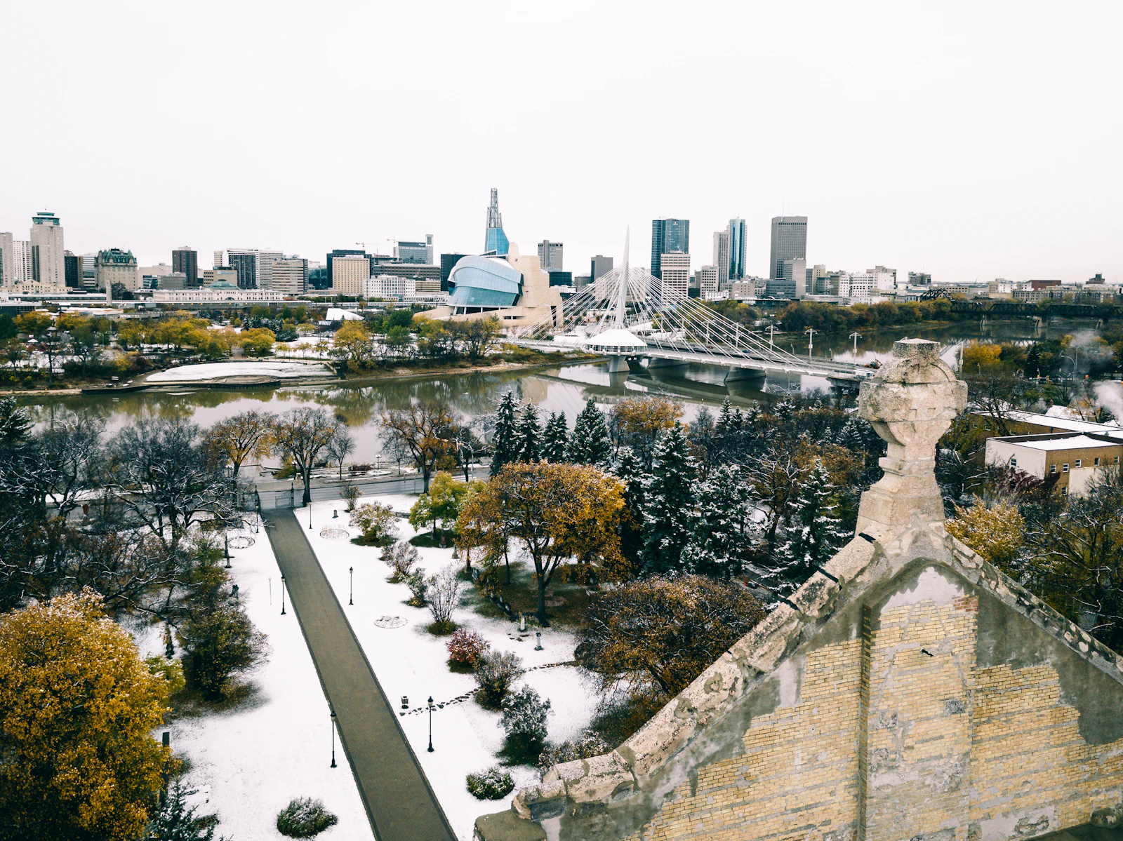 Green trees in the foreground with Winnipeg city office buildings rising behind them under daylight