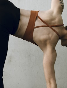 A person is in a yoga pose, showcasing their back and arm muscles. They are wearing a brown top and black pants. A tattoo is visible on their upper arm.