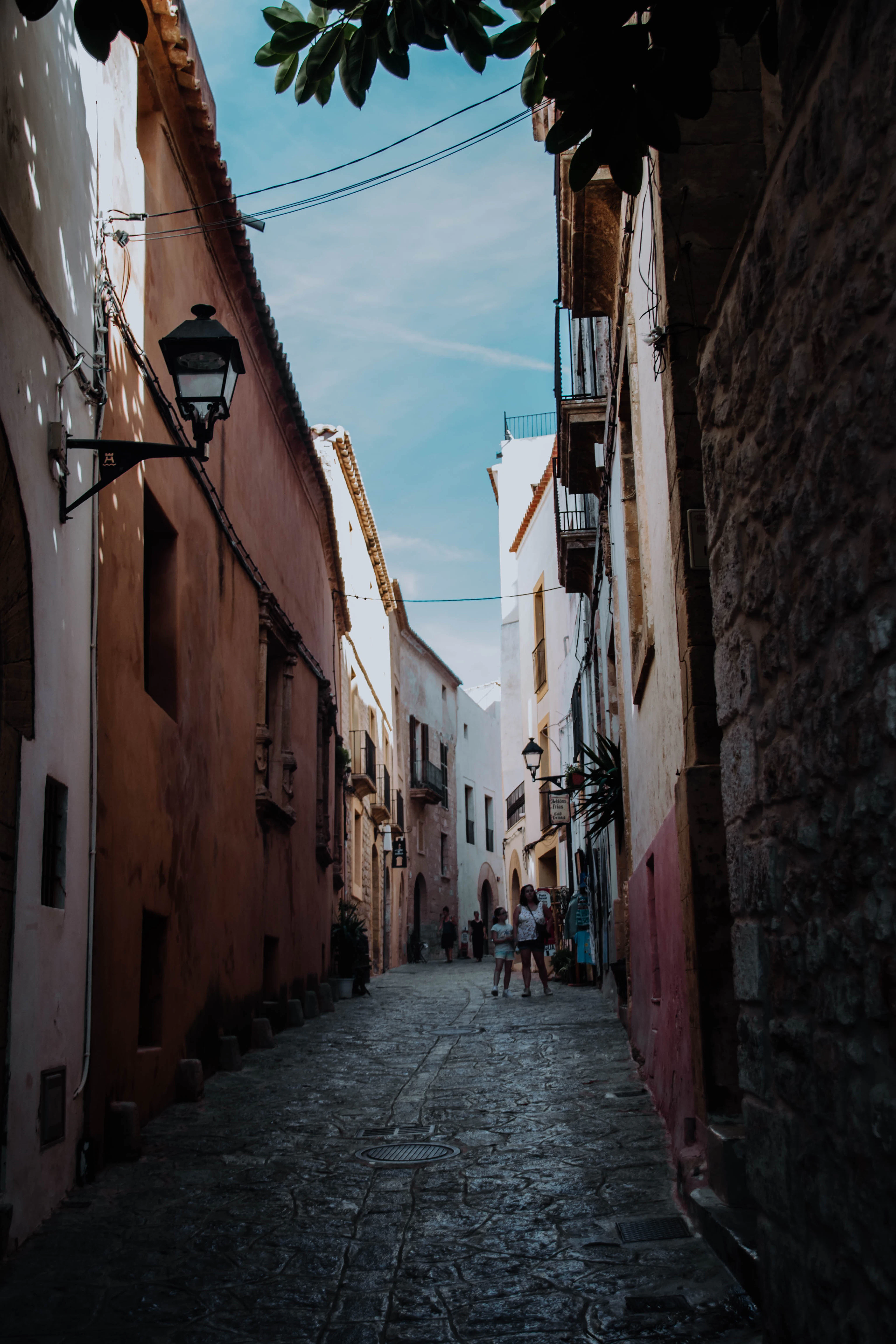 A quiet, shaded cobbled alley in Ibiza's old town, hemmed by ochre and pink stone walls with an iron street lantern and two figures at the sunlit far end
