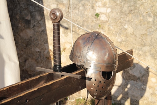 A medieval helmet and sword rest on a wooden structure against a stone wall. The helmet is metallic with rivets and detailed craftsmanship, while the sword has a simple, sturdy handle and blade.