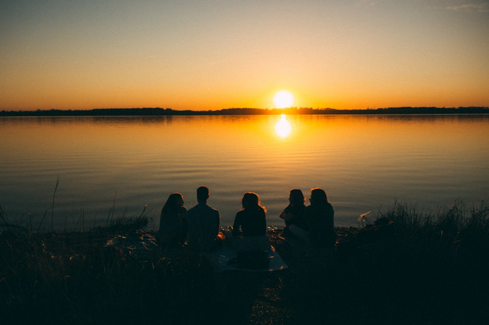 a group of people sitting on the shore of a lake at sunset