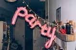 A festive arrangement in a dim hallway with pink balloon letters spelling 'party' is hanging from a string of small golden lights. The wall has a shelf with several bottles, a sign, and a small poster or picture. The area appears decorated for a celebration, with other miscellaneous items in the background adding to the party atmosphere.