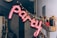 A festive arrangement in a dim hallway with pink balloon letters spelling 'party' is hanging from a string of small golden lights. The wall has a shelf with several bottles, a sign, and a small poster or picture. The area appears decorated for a celebration, with other miscellaneous items in the background adding to the party atmosphere.
