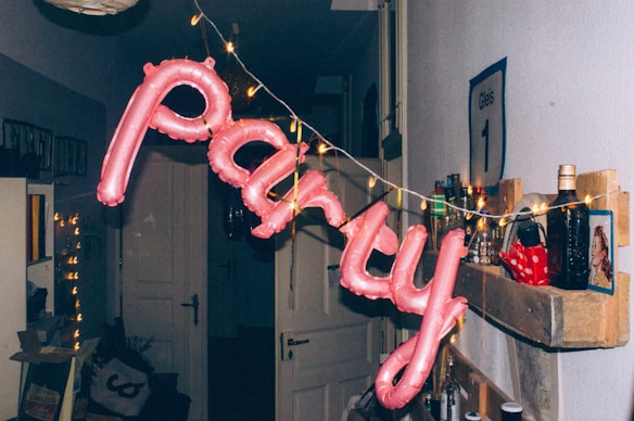 A festive arrangement in a dim hallway with pink balloon letters spelling 'party' is hanging from a string of small golden lights. The wall has a shelf with several bottles, a sign, and a small poster or picture. The area appears decorated for a celebration, with other miscellaneous items in the background adding to the party atmosphere.