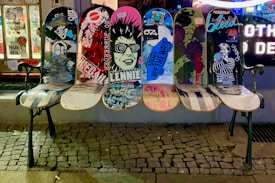 A bench made from repurposed skateboards with vibrant graphics and graffiti, positioned on a cobblestone sidewalk in front of a street art shop window. The boards display various artistic designs, including urban and punk imagery.