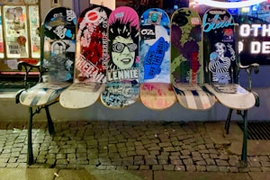 A bench made from repurposed skateboards with vibrant graphics and graffiti, positioned on a cobblestone sidewalk in front of a street art shop window. The boards display various artistic designs, including urban and punk imagery.