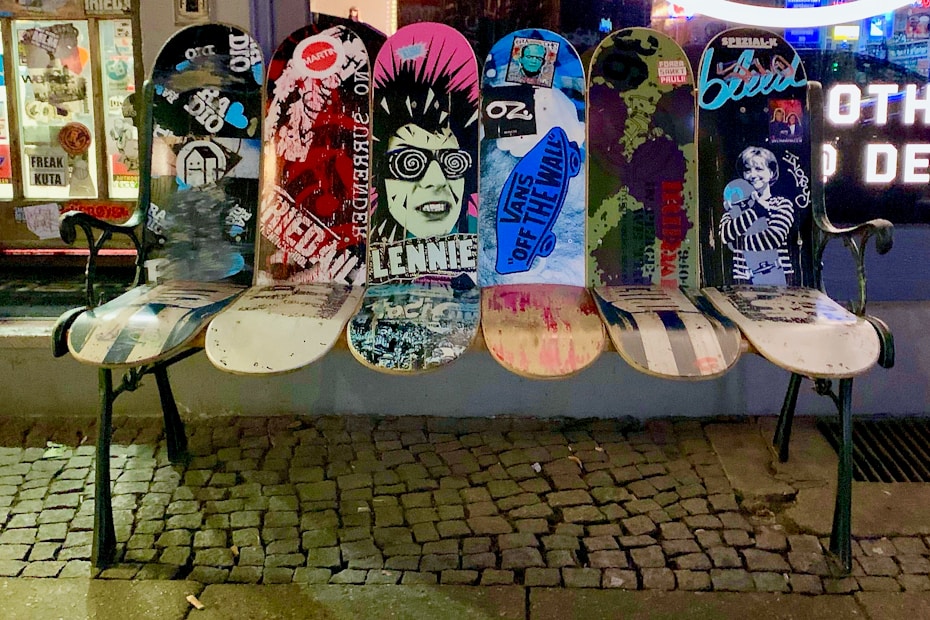 A bench made from repurposed skateboards with vibrant graphics and graffiti, positioned on a cobblestone sidewalk in front of a street art shop window. The boards display various artistic designs, including urban and punk imagery.