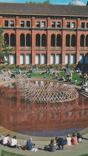 A courtyard features a large circular pool with a distinctive woven metal sculpture at its center, surrounded by people sitting on the edge of the pool and the grass. The backdrop is an impressive brick building with tall arched windows and ornate architectural details.