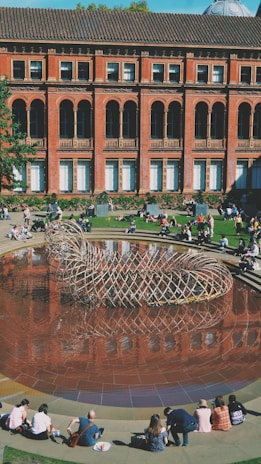 A courtyard features a large circular pool with a distinctive woven metal sculpture at its center, surrounded by people sitting on the edge of the pool and the grass. The backdrop is an impressive brick building with tall arched windows and ornate architectural details.