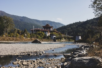 A serene landscape featuring a traditional Bhutanese architecture structure nestled among lush greenery, with a clear river flowing in the foreground and surrounded by forested mountains in the background.