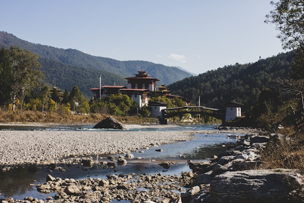 A serene landscape featuring a traditional Bhutanese architecture structure nestled among lush greenery, with a clear river flowing in the foreground and surrounded by forested mountains in the background.