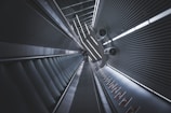 A futuristic-looking escalator in a modern building, viewed from a low angle. The scene is characterized by metallic surfaces and sleek, parallel lines. Two circular ventilation ducts extend from the ceiling, adding to the industrial aesthetic. Advertisements are visible on the right side wall.