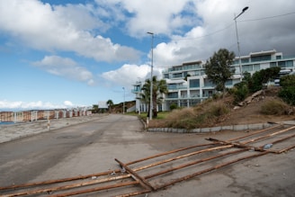 A scene showing a modern residential complex with multiple white buildings. The road leading to the complex is partially blocked by rusted metal barriers lying on the ground. There are trees and shrubs lining the pathway, and a clear blue sky with scattered clouds provides a contrast to the buildings. Street lamps are visible along the road.