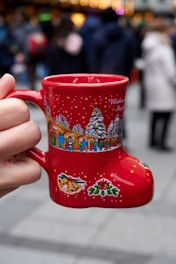 A red boot-shaped mug is being held by a hand, featuring festive designs such as a Christmas tree and colorful illustrations of people and holiday decorations. The background is blurred, showing a crowd of people in winter clothing, suggesting a busy, outdoor festive event.
