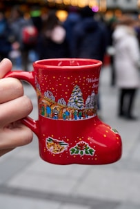 A red boot-shaped mug is being held by a hand, featuring festive designs such as a Christmas tree and colorful illustrations of people and holiday decorations. The background is blurred, showing a crowd of people in winter clothing, suggesting a busy, outdoor festive event.