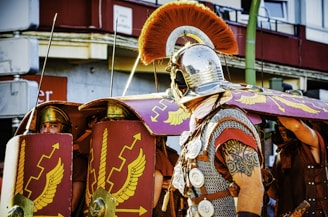 A group of people dressed in Roman legionary costumes, carrying large shields adorned with yellow winged designs and wielding helmets. The man in the foreground has a decorative metal helmet with a red crest and intricate chainmail armor. In the background, others form a protective formation using their shields.