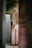 An old, abandoned bathroom with peeling walls and exposed plumbing is seen. The tiles are mostly pink, contrasting with a moldy, deteriorating green wall. The floor is dirty, and there is a general sense of neglect and decay.