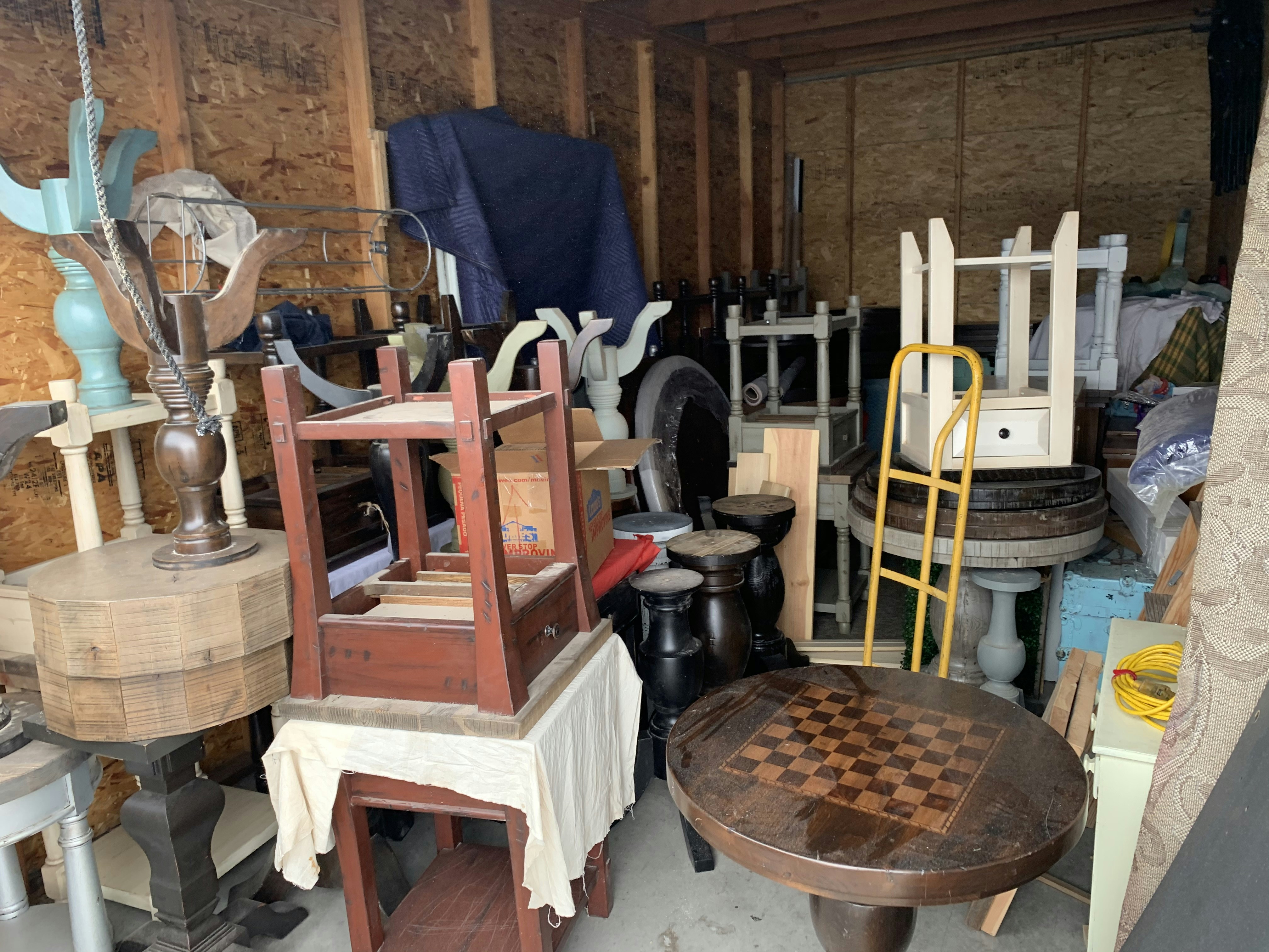 A cluttered storage area filled with various pieces of furniture and household items. Items include a checkerboard table, assorted chairs, stools, a hand truck, and other disassembled wooden furniture. The walls are unfinished, with exposed beams, creating a rustic backdrop.