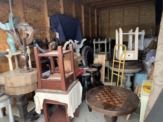 A cluttered storage area filled with various pieces of furniture and household items. Items include a checkerboard table, assorted chairs, stools, a hand truck, and other disassembled wooden furniture. The walls are unfinished, with exposed beams, creating a rustic backdrop.