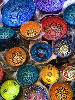 A collection of intricately designed decorative bowls with vibrant colors and patterns. The bowls feature floral and geometric designs, with each one displaying unique artistry and color combinations. They are stacked and nestled with packaging material around them.