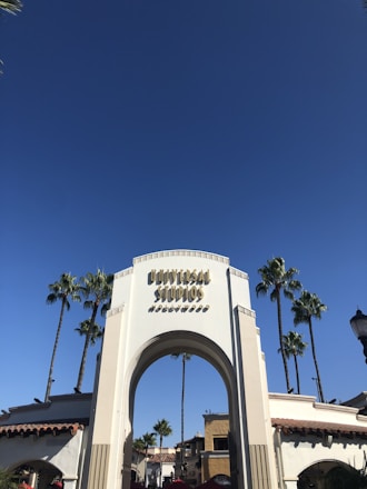 A large white archway with the words 'Universal Studios Hollywood' prominently displayed. Tall palm trees surround the structure under a clear blue sky. Buildings with red-tiled roofs are visible in the background.