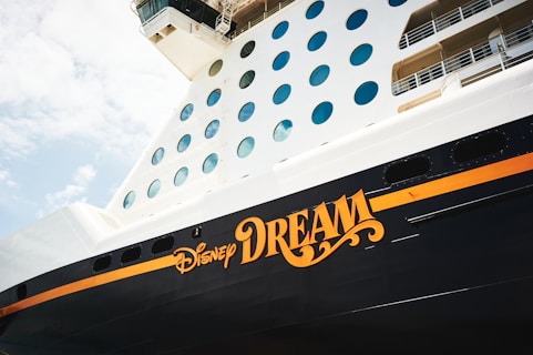 A close-up view of the side of a cruise ship featuring large circular windows and the words 'Disney Dream' prominently displayed in bold, stylized lettering. The ship's surface is painted in dark and light contrasting colors, creating a striking visual. The sky is visible in the background, giving a sense of openness and adventure.