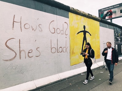 Graffiti on a wall displays the text 'How's God? She's black.' Next to the text is a yellow section featuring a black silhouette of a person. A man and a woman walk by, the woman looking at her phone.