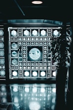 A decorative wall composed of intricate geometric patterns featuring illuminated panels with various shapes like circles and hexagons. A large circular window is the focal point, allowing light to stream through. A potted plant is visible on the right side, casting a shadow on the reflective floor.