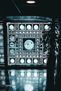 A decorative wall composed of intricate geometric patterns featuring illuminated panels with various shapes like circles and hexagons. A large circular window is the focal point, allowing light to stream through. A potted plant is visible on the right side, casting a shadow on the reflective floor.