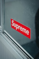 A reflective surface displays a red sticker with the brand name 'Supreme' in bold white letters. The sticker appears to be affixed onto a glass pane, with metallic or wooden trim visible along the edges.